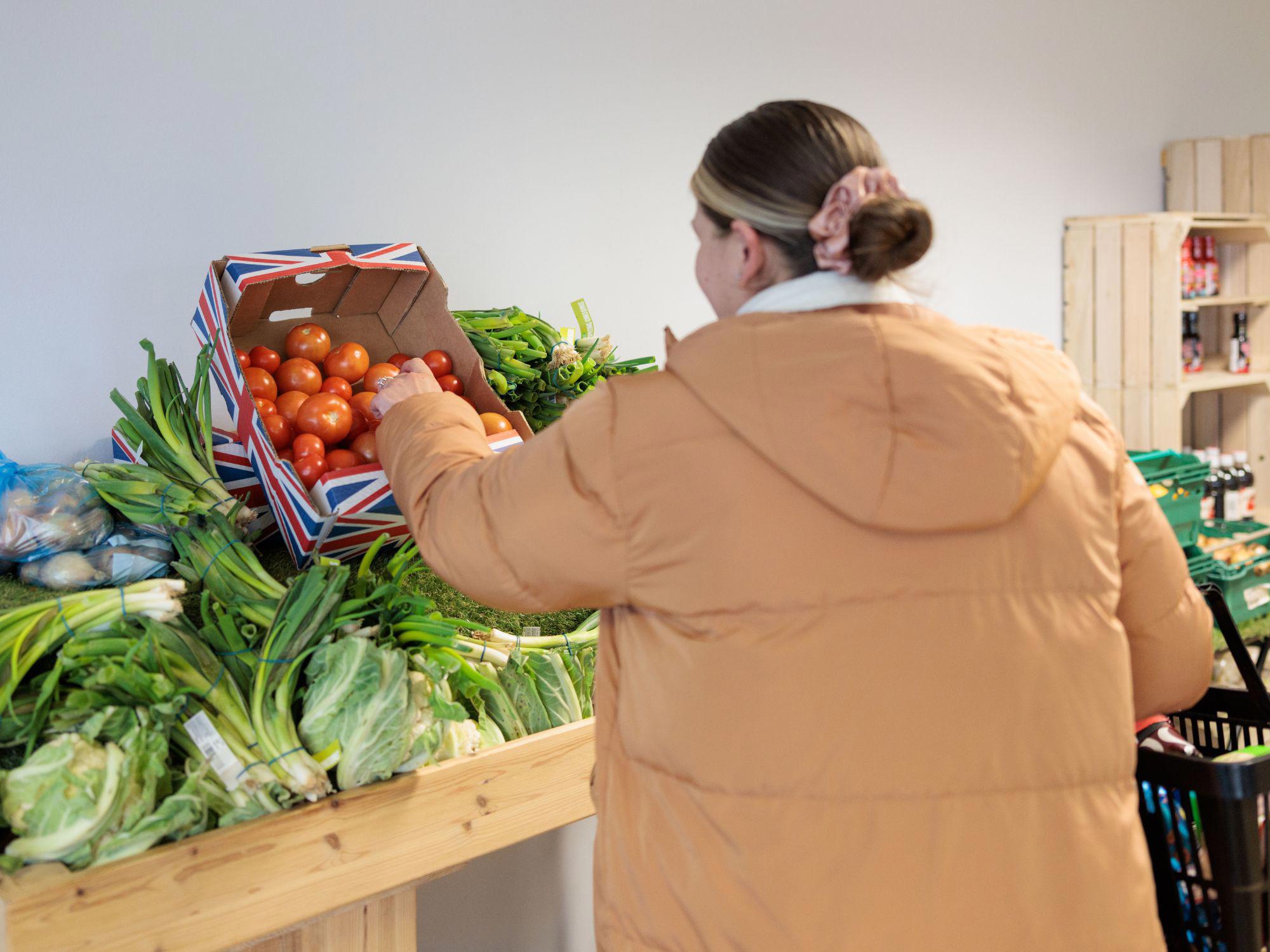 Person in a tan jacket selecting fresh tomatoes from a wooden table filled with assorted vegetables at The Market Place, with shelves of produce in the background.
