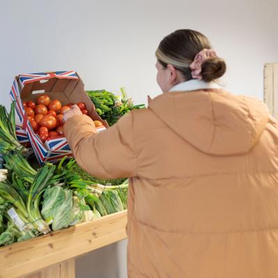 Person in a tan jacket selecting fresh tomatoes from a wooden table filled with assorted vegetables at The Market Place, with shelves of produce in the background.