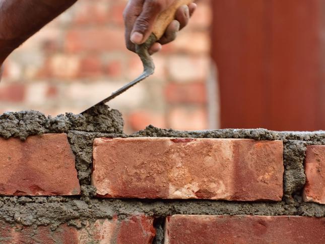 A close-up of a hand using a trowel to apply mortar between red bricks while constructing a brick wall. A close-up of a hand using a trowel to apply mortar between red bricks while constructing a brick wall.