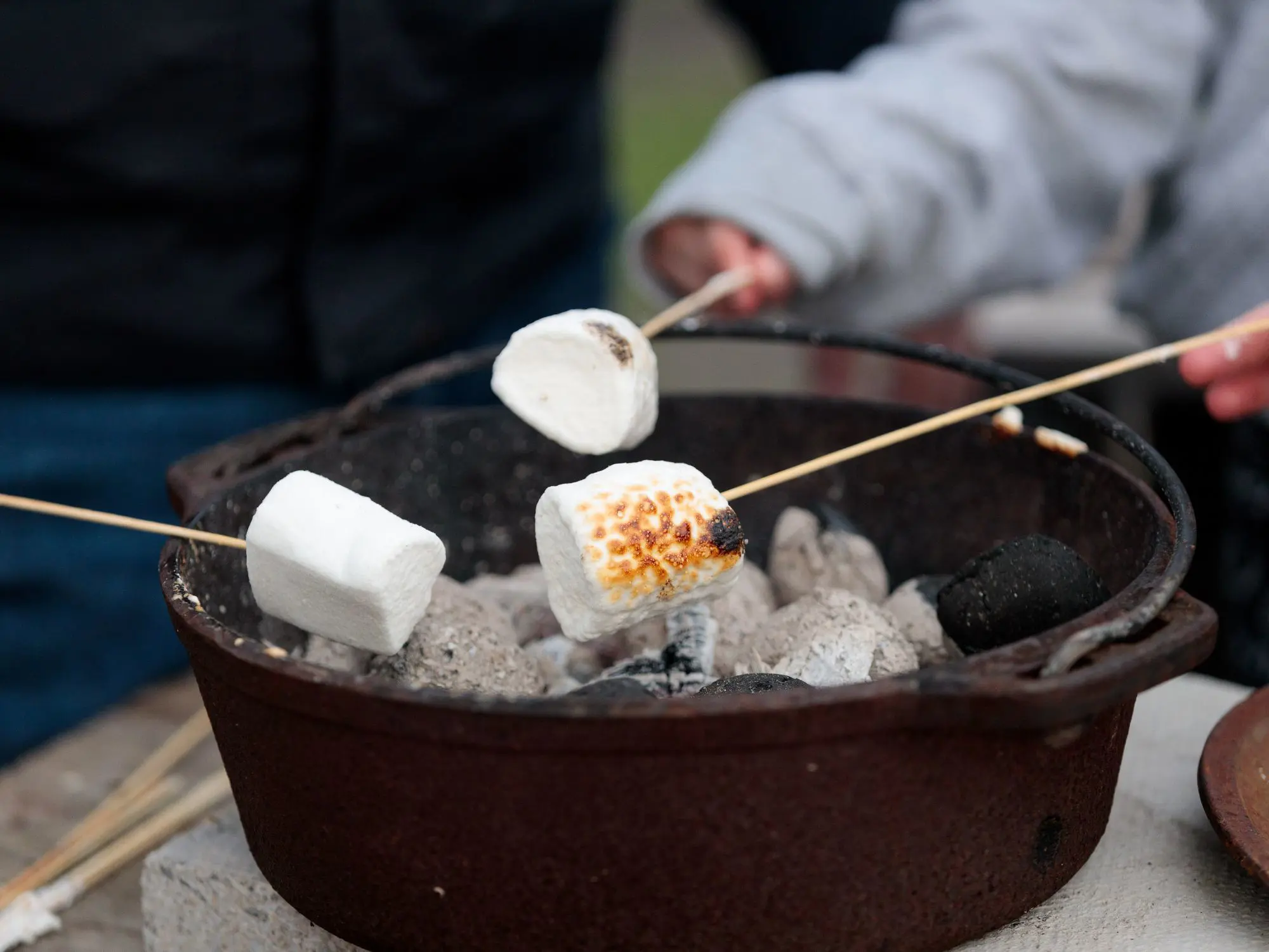 Three marshmallows on wooden sticks being roasted over a charcoal fire pit, with one marshmallow partially toasted to a golden-brown color, suggesting an outdoor bonfire or s'mores-making activity.