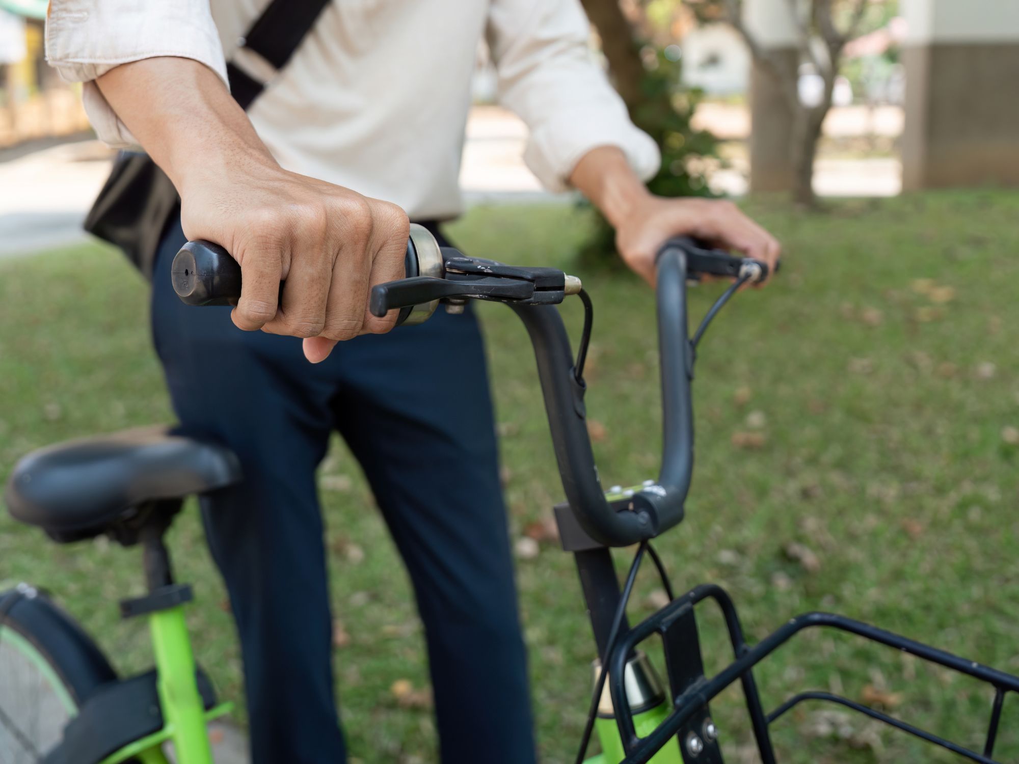 Person holding the handlebars of a green bicycle in an outdoor setting with grass and trees, wearing a light long-sleeve shirt and dark trousers