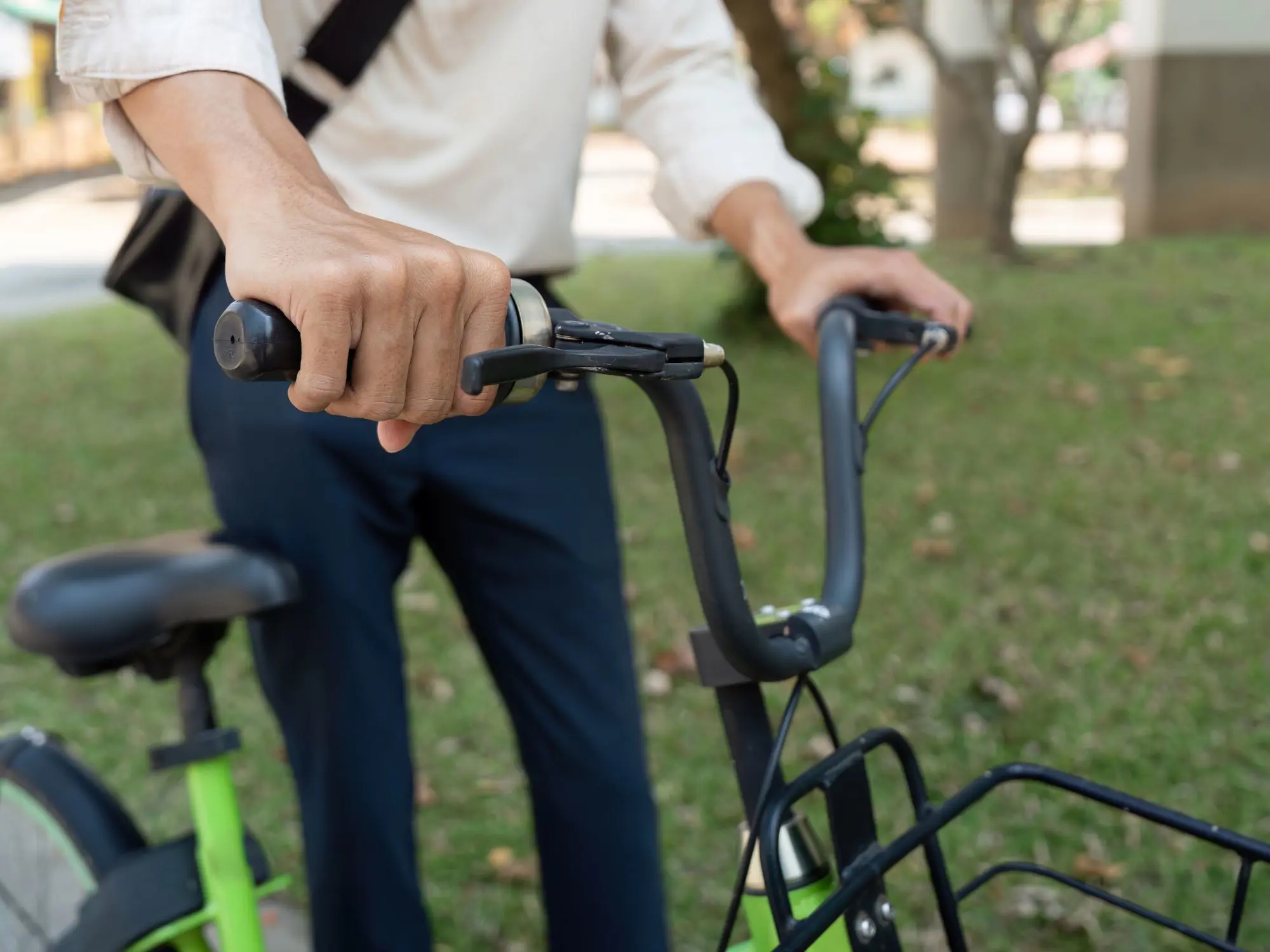 Person holding the handlebars of a green bicycle in an outdoor setting with grass and trees, wearing a light long-sleeve shirt and dark trousers
