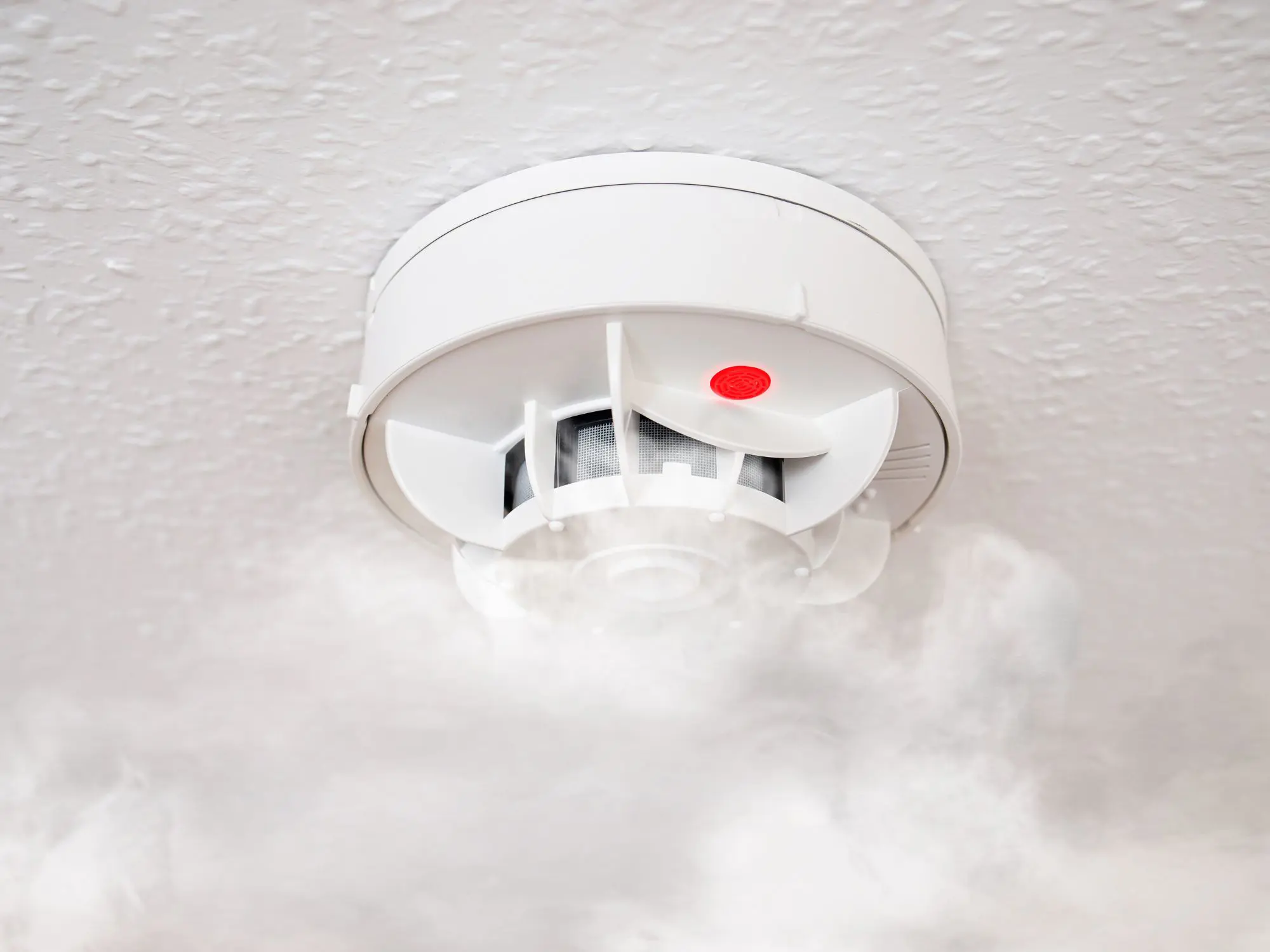 Smoke detector with red light activated and smoke rising around it.