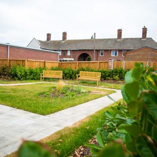 Communal landscaped garden with three wooden benches, planted borders, paved paths and neighbouring homes visible in the background.