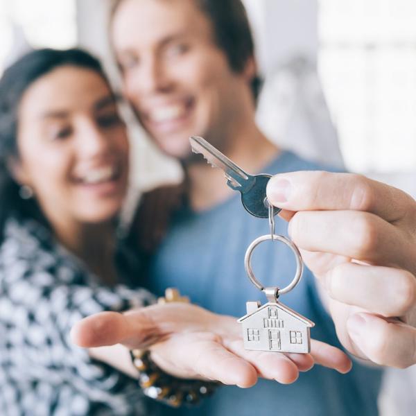 Two people holding a key with a house-shaped keychain; one person extends the key toward the camera while the other holds an open hand beneath it, indoors with natural light in the background.