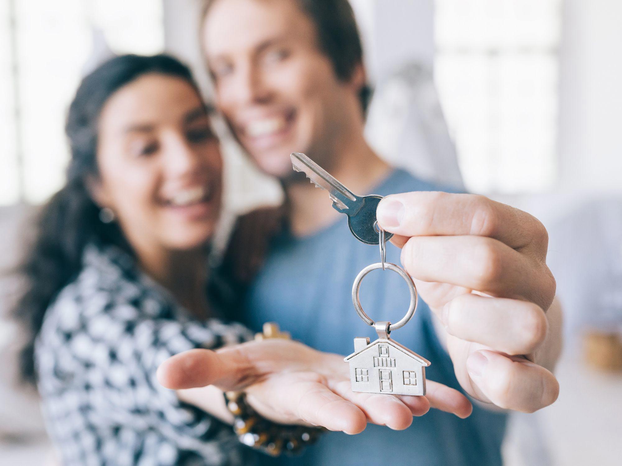 Two people holding a key with a house-shaped keychain; one person extends the key toward the camera while the other holds an open hand beneath it, indoors with natural light in the background.