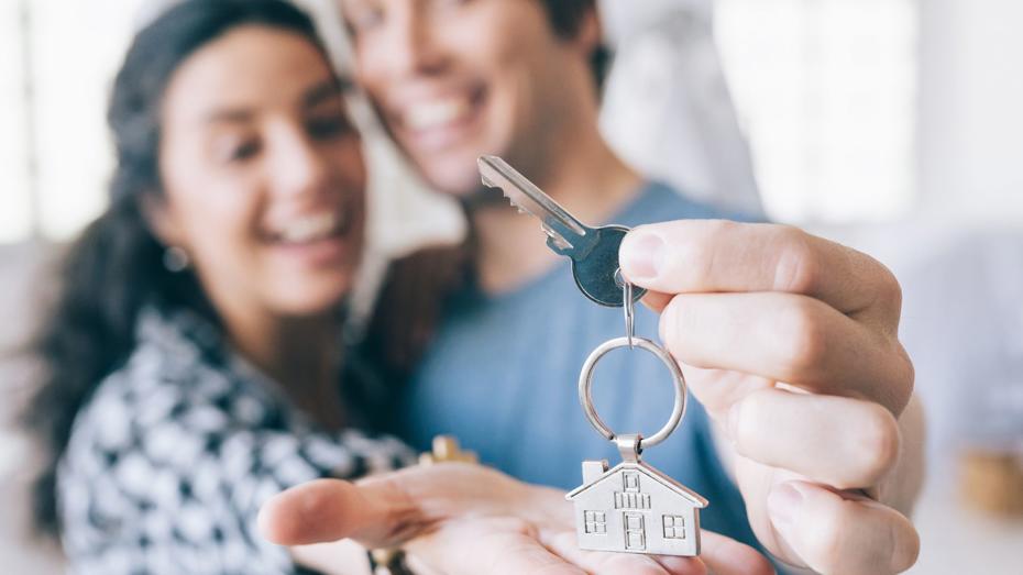 Two people holding a key with a house-shaped keychain; one person extends the key toward the camera while the other holds an open hand beneath it, indoors with natural light in the background. Two people holding a key with a house-shaped keychain; one person extends the key toward the camera while the other holds an open hand beneath it, indoors with natural light in the background.