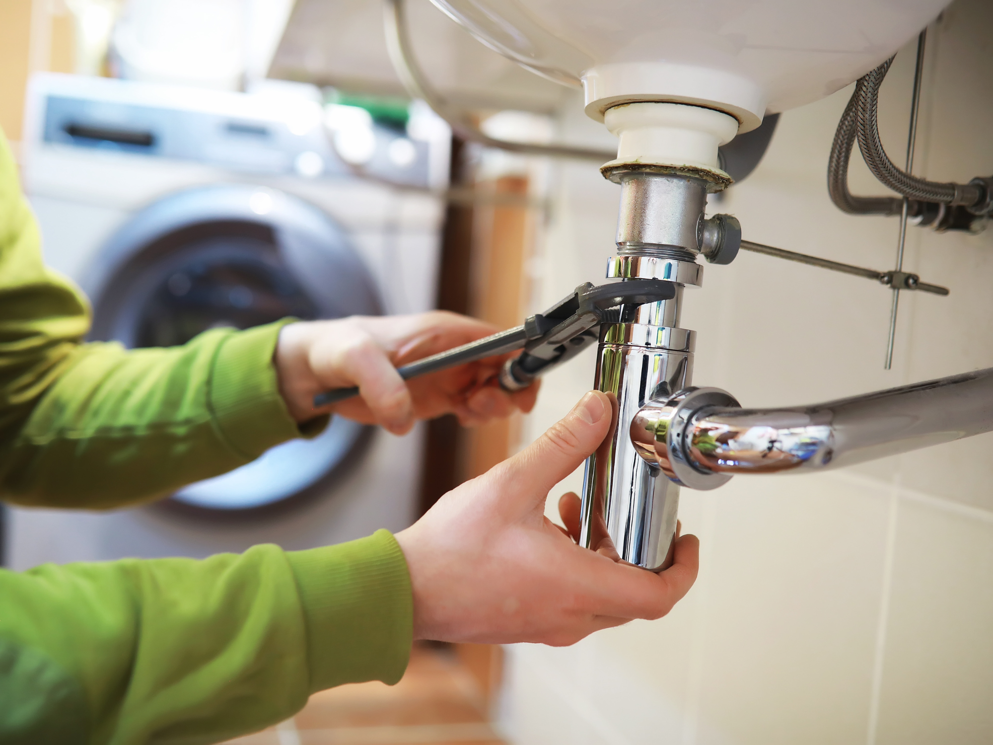 Hands using an adjustable wrench on plumbing fittings under a sink, with a washing machine in the background.