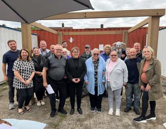Group of 18 people posing outdoors under a wooden structure, with a red shipping container in the background. Some individuals are holding papers, and a partially visible umbrella is in the top left corner. The group appears to be gathered for an event or meeting.