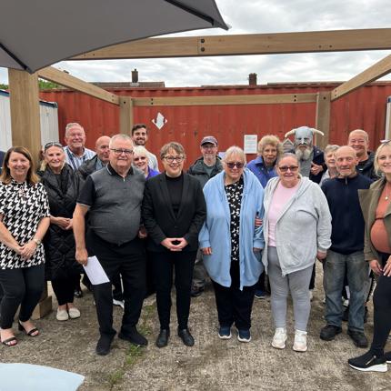 Group of 18 people posing outdoors under a wooden structure, with a red shipping container in the background. Some individuals are holding papers, and a partially visible umbrella is in the top left corner. The group appears to be gathered for an event or meeting.