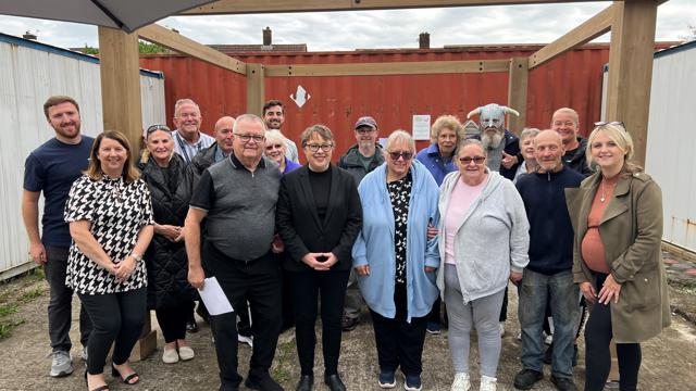 Group of 18 people posing outdoors under a wooden structure, with a red shipping container in the background. Some individuals are holding papers, and a partially visible umbrella is in the top left corner. The group appears to be gathered for an event or meeting.