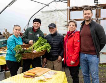 “A group of people standing together inside a greenhouse, with two individuals holding a large basket filled with freshly harvested vegetables. A table in front of them is covered with a yellow cloth and displays various vegetables and food items. Gardening and cooking equipment can be seen in the background.”