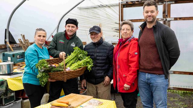 “A group of people standing together inside a greenhouse, with two individuals holding a large basket filled with freshly harvested vegetables. A table in front of them is covered with a yellow cloth and displays various vegetables and food items. Gardening and cooking equipment can be seen in the background.”