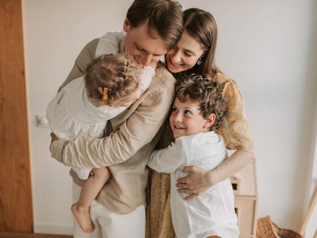 Two adults embracing two young children in a warm, home setting, with one child being held and the other hugging from the front. Two adults embracing two young children in a warm, home setting, with one child being held and the other hugging from the front.