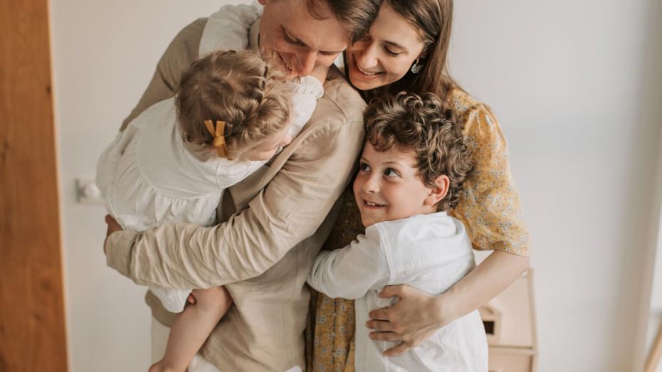 Two adults embracing two young children in a warm, home setting, with one child being held and the other hugging from the front. Two adults embracing two young children in a warm, home setting, with one child being held and the other hugging from the front.