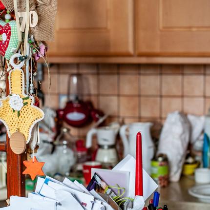 Cluttered kitchen with various appliances and utensils on the counter. In the foreground, a wooden structure displays hanging decorations including a heart, a cross with a flower, and an orange star. A container filled with papers and envelopes is also visible.