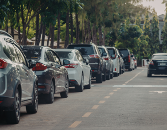 A row of parked cars lines one side of a residential street bordered by trees and greenery. Vehicles are parked closely together along the kerb, while the road ahead remains clear for traffic. The image is taken from street level, showing the length of the parked cars stretching into the distance.