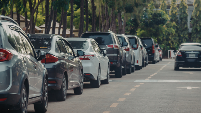 A row of parked cars lines one side of a residential street bordered by trees and greenery. Vehicles are parked closely together along the kerb, while the road ahead remains clear for traffic. The image is taken from street level, showing the length of the parked cars stretching into the distance.