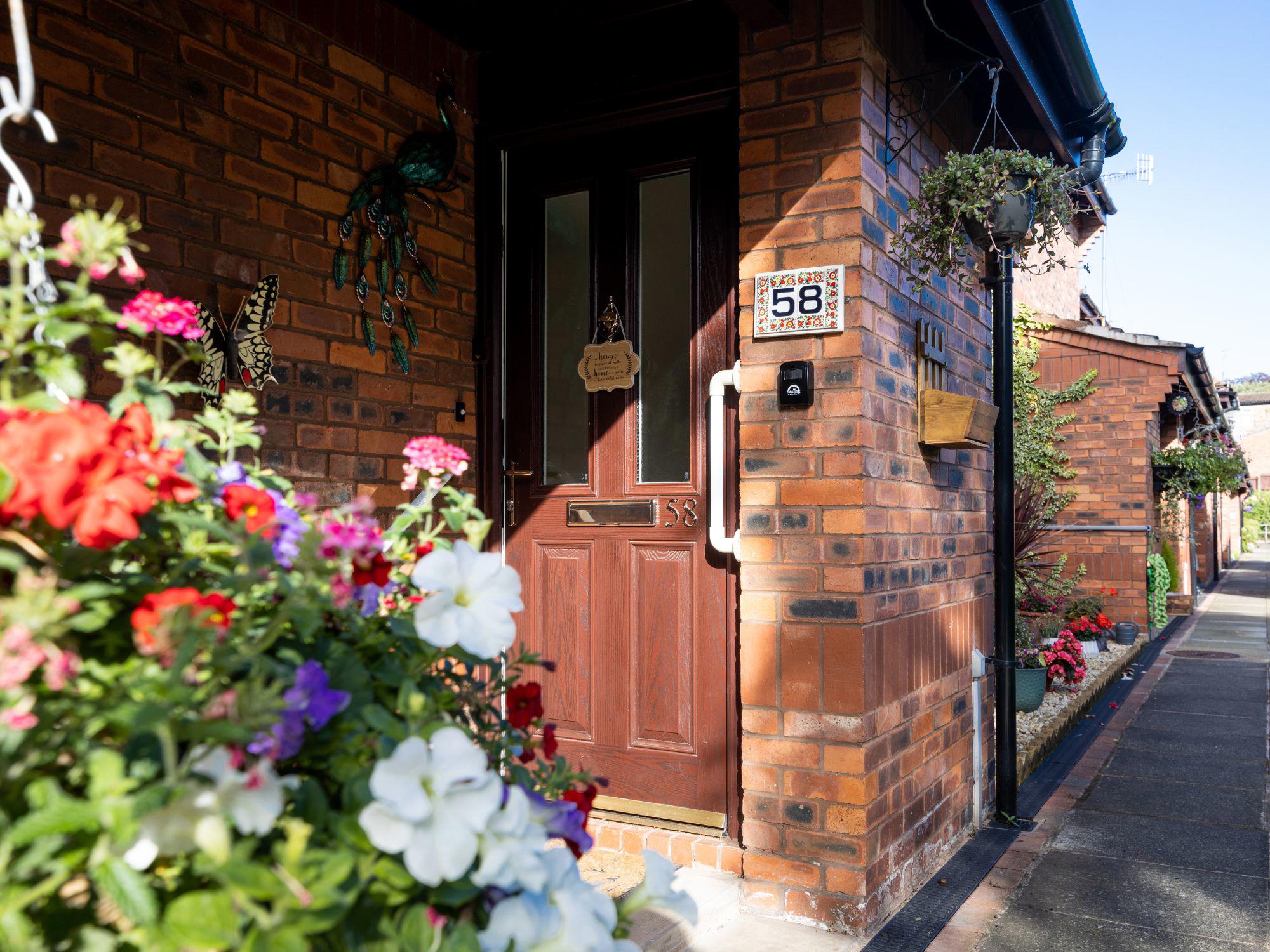 Entrance to a brick house with a wooden door marked '58', surrounded by colourful flowers in hanging baskets and pots. Similar houses line the pathway.