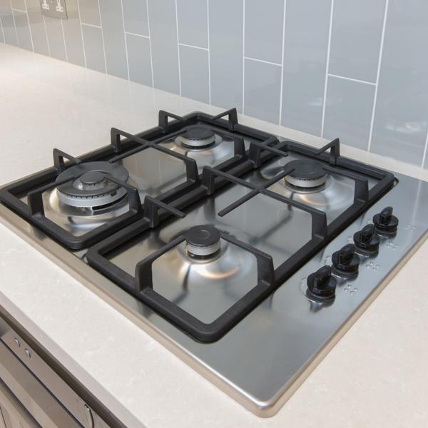 Modern stainless steel gas cooktop with four burners and black grates, set into a light-coloured countertop with grey tiled backsplash and control knobs on the right.