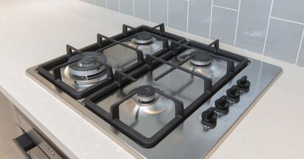 Modern stainless steel gas cooktop with four burners and black grates, set into a light-coloured countertop with grey tiled backsplash and control knobs on the right.