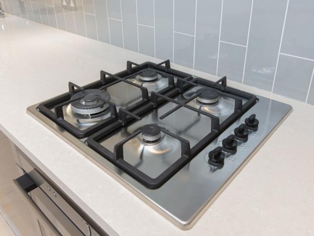 Modern stainless steel gas cooktop with four burners and black grates, set into a light-coloured countertop with grey tiled backsplash and control knobs on the right. Modern stainless steel gas cooktop with four burners and black grates, set into a light-coloured countertop with grey tiled backsplash and control knobs on the right.