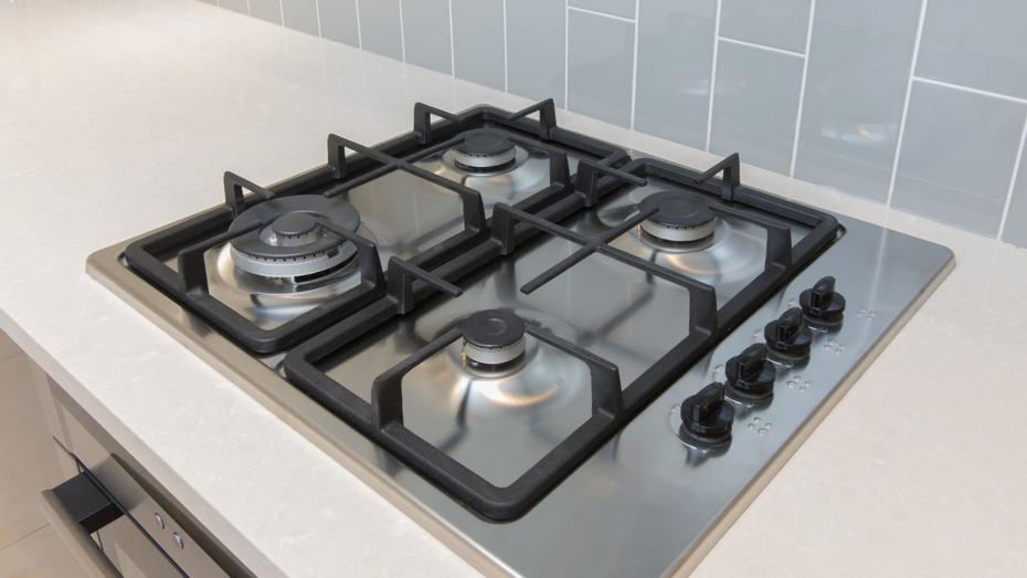 Modern stainless steel gas cooktop with four burners and black grates, set into a light-coloured countertop with grey tiled backsplash and control knobs on the right. Modern stainless steel gas cooktop with four burners and black grates, set into a light-coloured countertop with grey tiled backsplash and control knobs on the right.