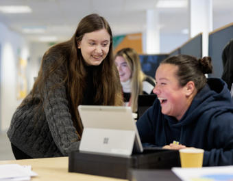 Two individuals smiling and collaborating over a laptop in an office setting, with papers and a cup on the desk. A third person is visible in the background working at a computer.