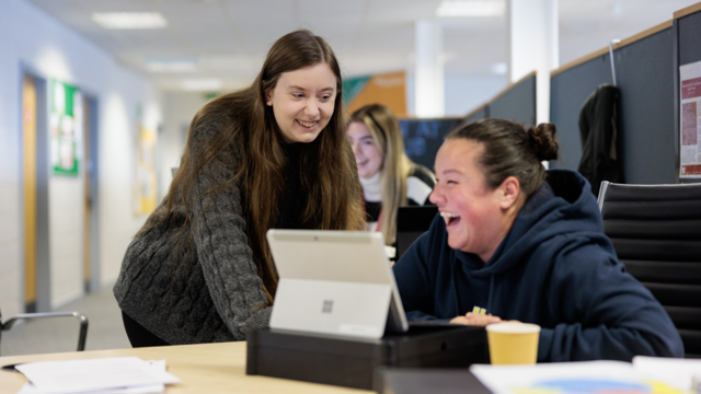 Two individuals smiling and collaborating over a laptop in an office setting, with papers and a cup on the desk. A third person is visible in the background working at a computer.