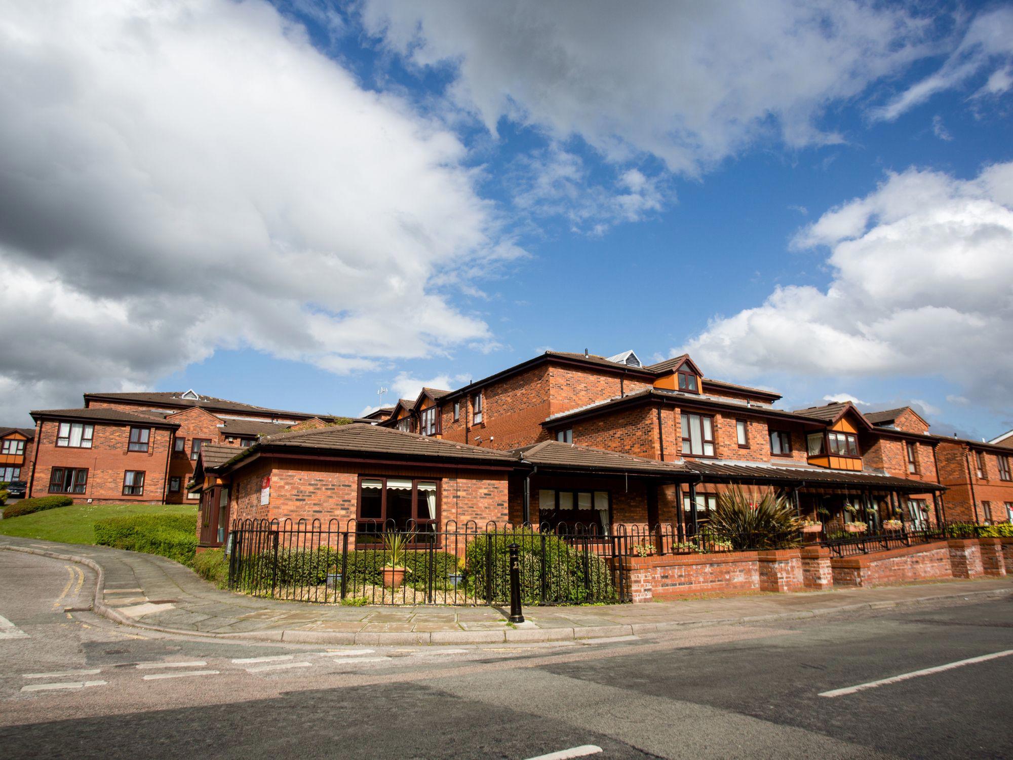 Residential area with brick buildings, brown roofs, and surrounding greenery under a partly cloudy sky. A black metal fence borders the property, with roads and a pedestrian crossing in the foreground.