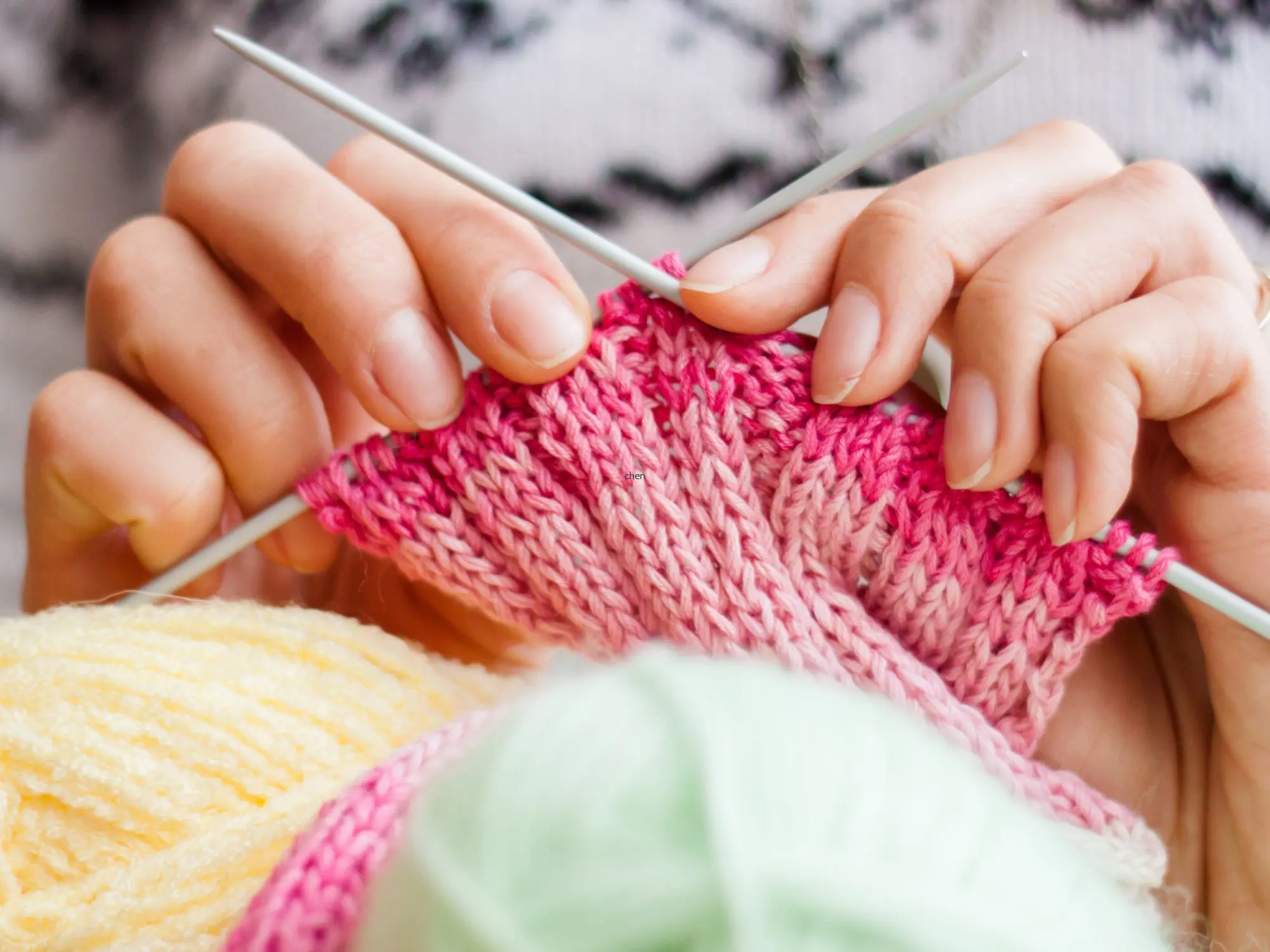 Close-up of hands knitting with pink yarn using two needles, with balls of yellow and mint green yarn in the foreground.