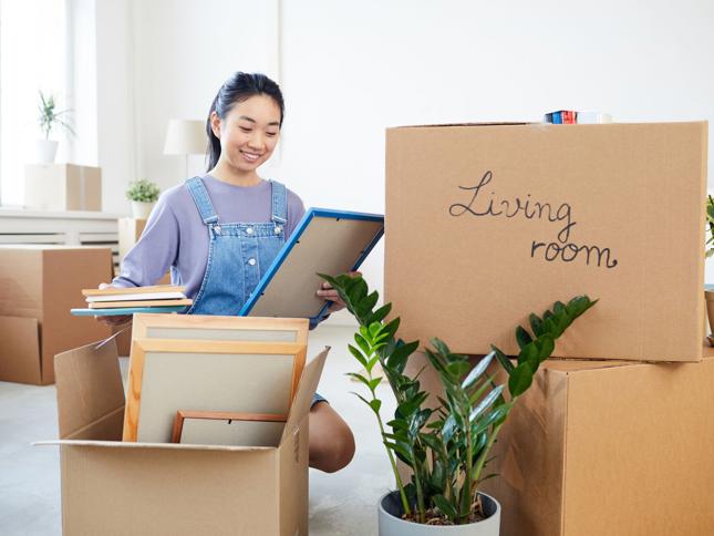 Person unpacking framed pictures and books from cardboard boxes in a bright living room, with a box labelled Living room and a green houseplant nearby. Person unpacking framed pictures and books from cardboard boxes in a bright living room, with a box labelled Living room and a green houseplant nearby.