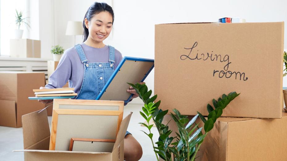 Person unpacking framed pictures and books from cardboard boxes in a bright living room, with a box labelled Living room and a green houseplant nearby. Person unpacking framed pictures and books from cardboard boxes in a bright living room, with a box labelled Living room and a green houseplant nearby.