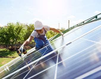 Person wearing a hard hat and gloves using a power drill to install solar panels on a rooftop, with trees and a building visible in the background.