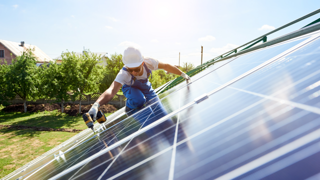 Person wearing a hard hat and gloves using a power drill to install solar panels on a rooftop, with trees and a building visible in the background.