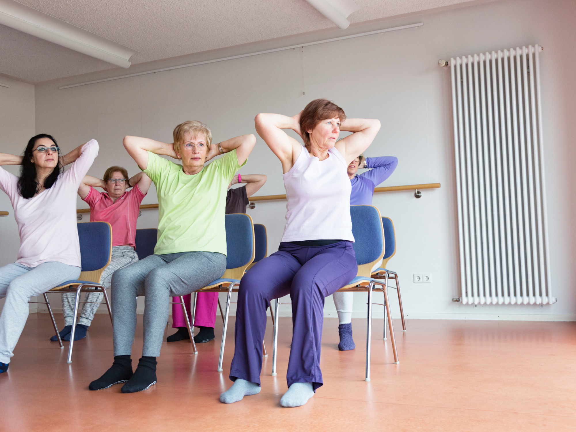 A group of people seated on chairs in a bright room, taking part in a gentle exercise session with their hands resting behind their heads, as part of a seated movement or wellbeing class.