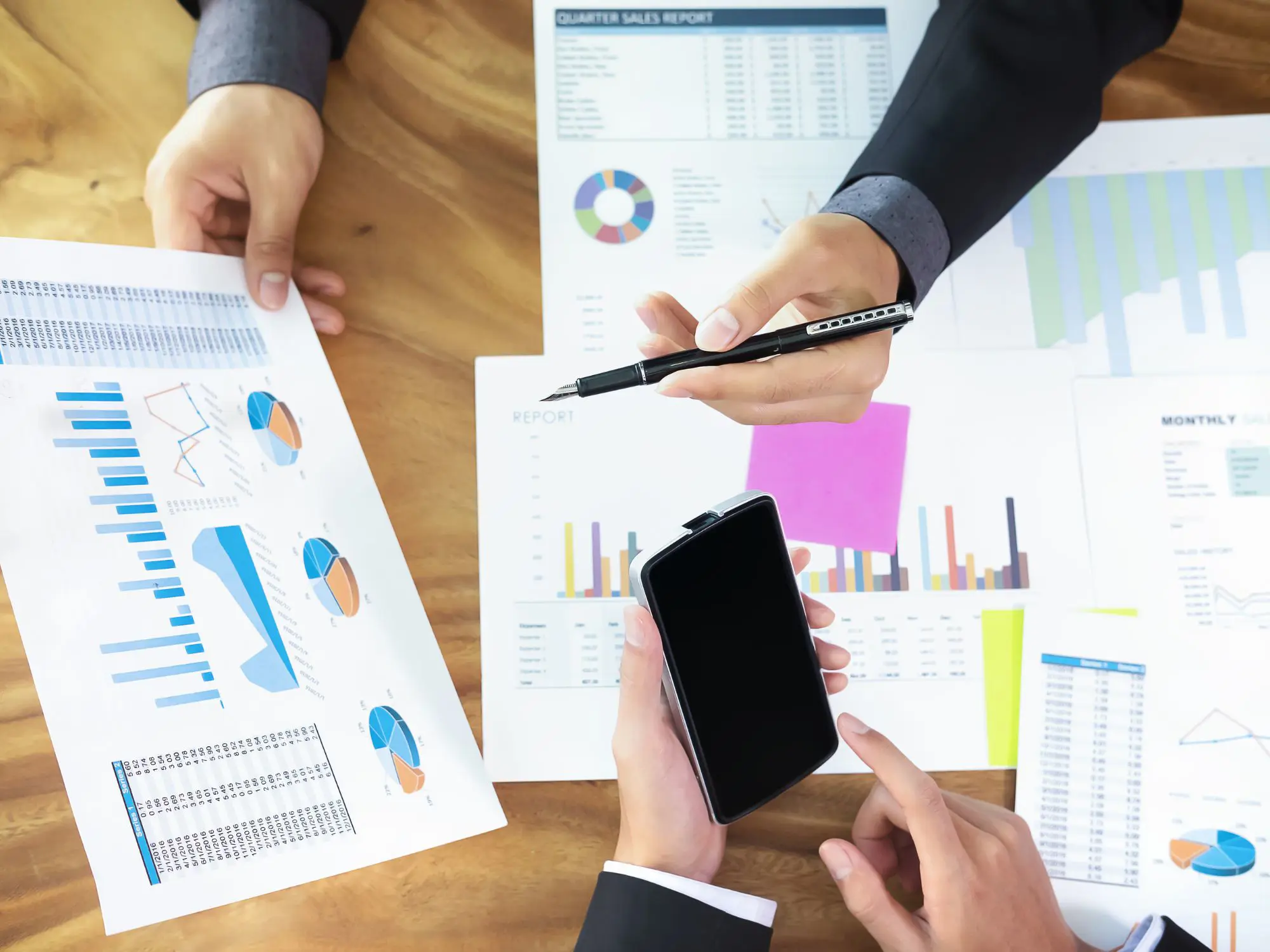 Three people in business attire sitting around a wooden table, reviewing printed charts and graphs including bar, pie, and line graphs. One person points with a pen, another holds a smartphone.