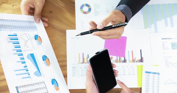 Three people in business attire sitting around a wooden table, reviewing printed charts and graphs including bar, pie, and line graphs. One person points with a pen, another holds a smartphone.