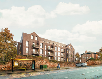 A street‑level view of a modern red‑brick apartment building, seen from across the road near a yellow bus shelter. The stepped roofline, balconies, and large windows are visible above a low sandstone wall. A couple of people walk along the pavement, and a parked car sits near the wall. Trees with autumn leaves frame the scene against a pale, cloudy sky.