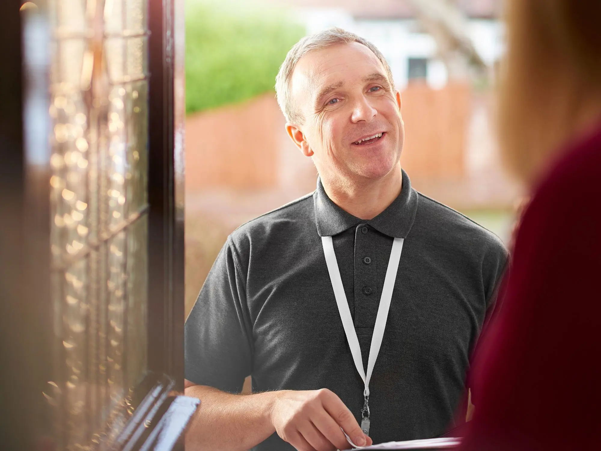 A person wearing a dark polo shirt and lanyard stands at a doorway, speaking with someone inside. The background shows greenery and residential buildings—suggesting a professional home visit or customer interaction.