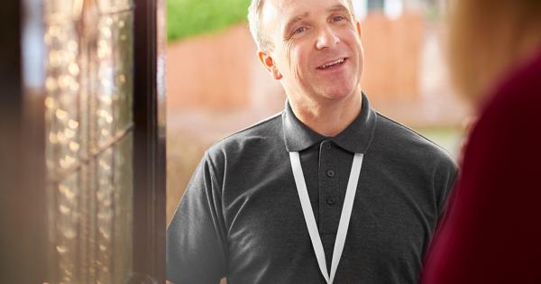 A person wearing a dark polo shirt and lanyard stands at a doorway, speaking with someone inside. The background shows greenery and residential buildings—suggesting a professional home visit or customer interaction.