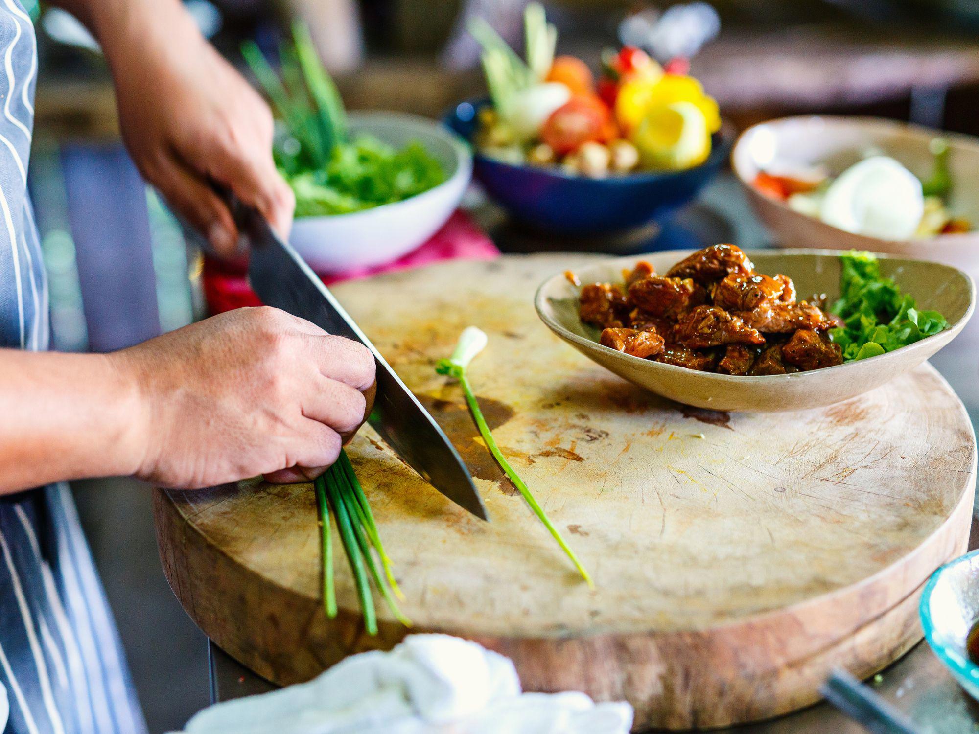 Close-up of hands chopping green onions on a wooden board, with a bowl of cooked food and fresh vegetables in the background.