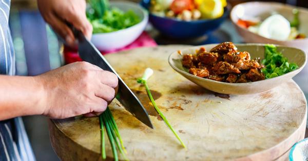 Close-up of hands chopping green onions on a wooden board, with a bowl of cooked food and fresh vegetables in the background.