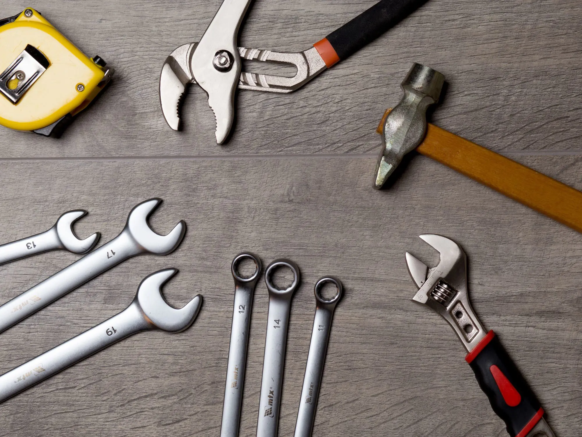 A variety of hand tools arranged on a wooden surface, including a yellow tape measure, two adjustable wrenches with orange and red handles, a yellow-handled hammer, and several open-end and combination wrenches labeled with sizes ranging from 10 to 17.