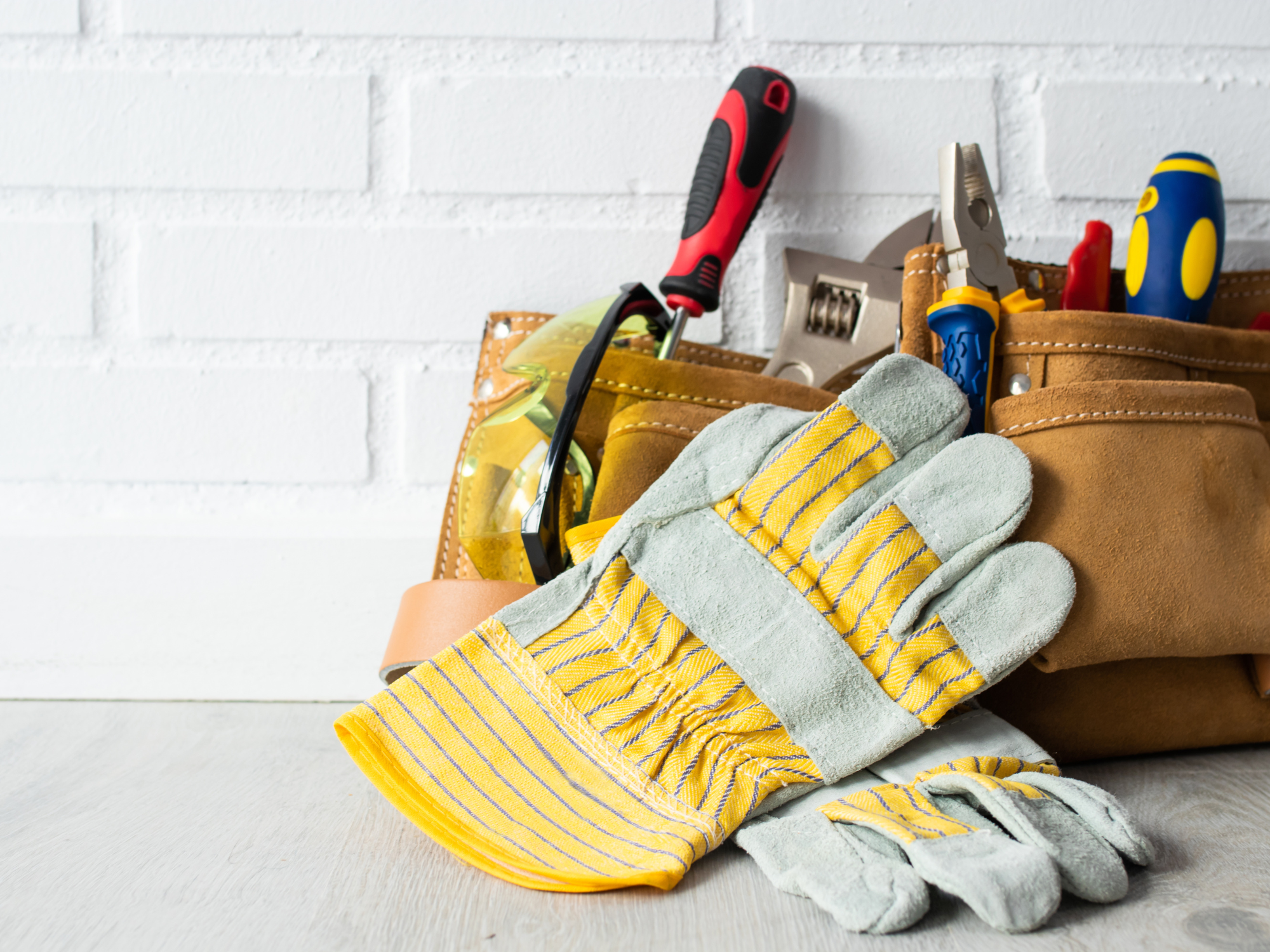  Tool belt filled with screwdrivers, pliers, and safety glasses, with yellow-striped work gloves in front.