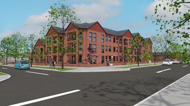 Street corner with a large three-story brick apartment building, surrounded by trees and sidewalks. Several people are walking and cars are visible on the road under a partly cloudy blue sky.