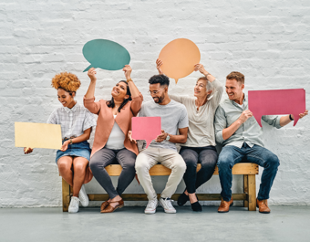 “Six people sitting on a wooden bench against a white brick wall, each holding colorful speech bubble or rectangular cut-out signs in various shapes and sizes, suggesting conversation or ideas.”