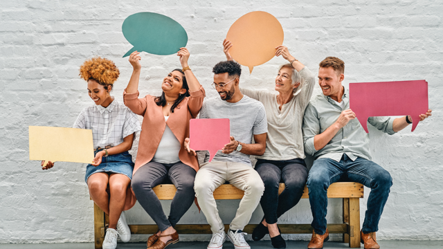 “Six people sitting on a wooden bench against a white brick wall, each holding colorful speech bubble or rectangular cut-out signs in various shapes and sizes, suggesting conversation or ideas.”
