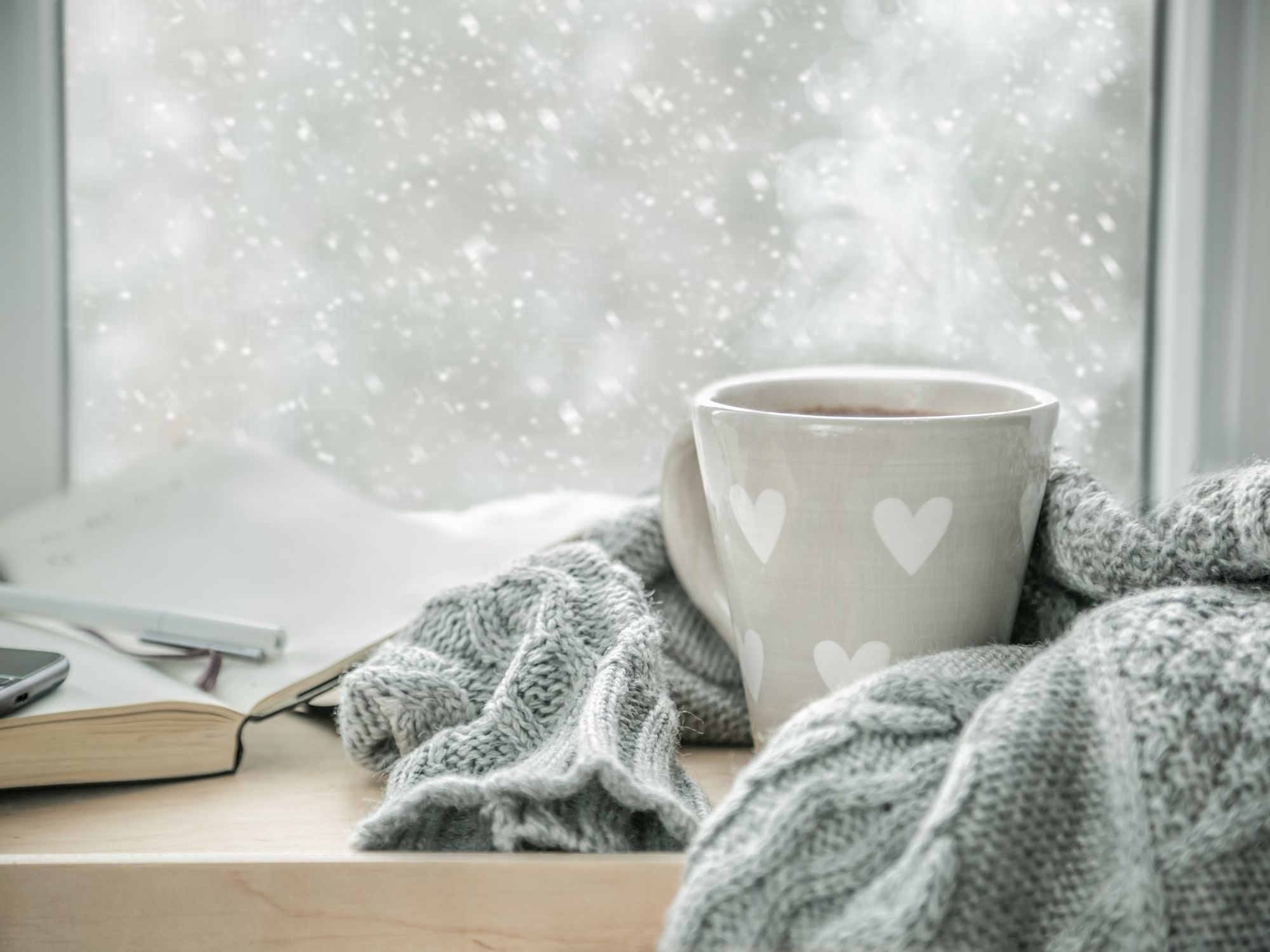 A steaming mug with white heart designs sits on a wooden surface wrapped in a gray knitted scarf, with an open book, pen, and smartphone nearby. Snow falls outside the window, creating a cozy winter scene.