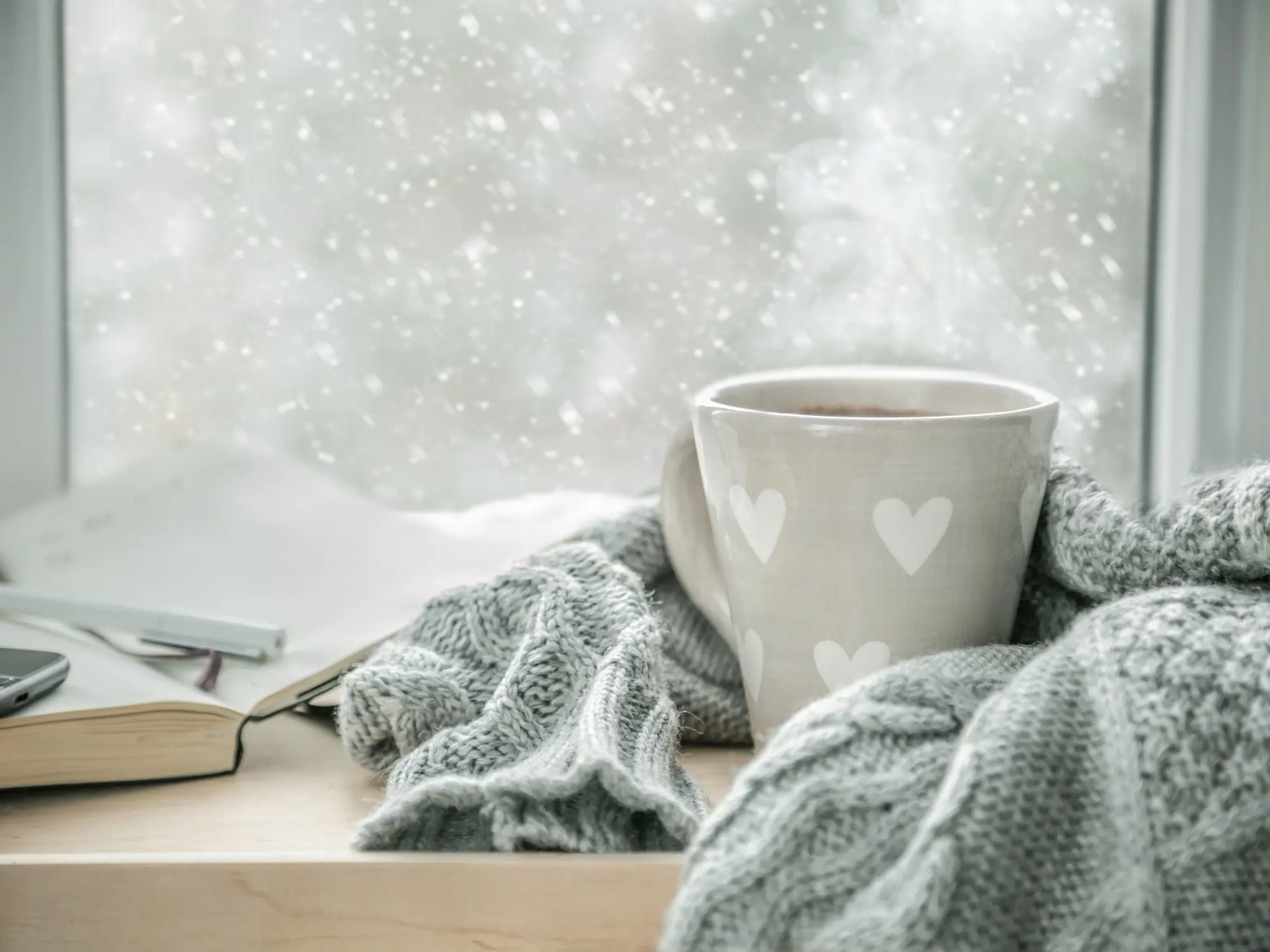 A steaming mug with white heart designs sits on a wooden surface wrapped in a gray knitted scarf, with an open book, pen, and smartphone nearby. Snow falls outside the window, creating a cozy winter scene.